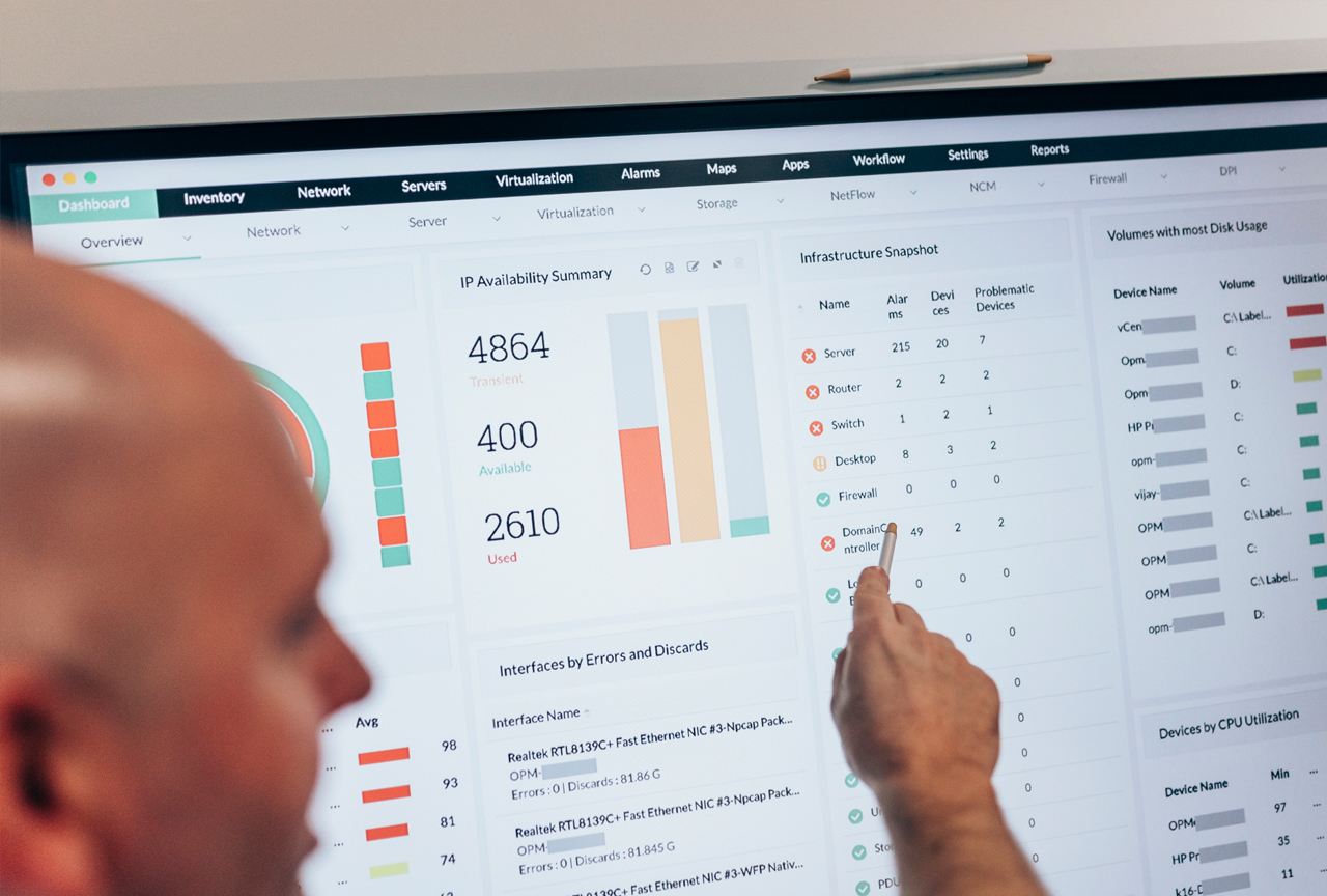 Proaktive Systemüberwachung: ein Ansprechpartner prüft Server, Netzwerk und Backup IT-Administrator zeigt mit einem Stift auf ein Monitoring-Dashboard mit Servern, Routern und Firewalls